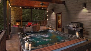 A wooden deck with a hot tub, wicker furniture, a stone fireplace, and a grill.