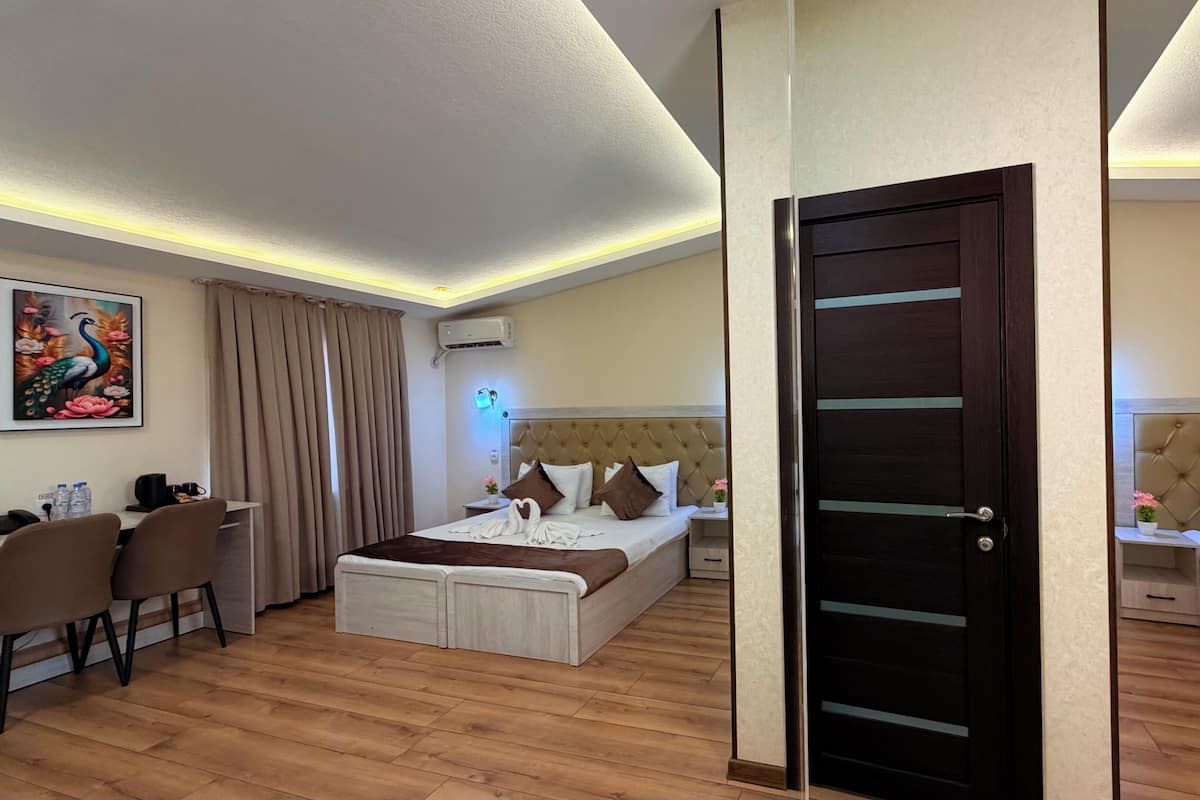 A hotel room with a bed, a desk, a chair, a door, and a painting on the wall.