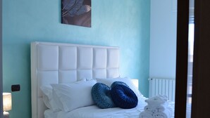 Una cama bien hecha con ropa blanca y almohadas azules, una mesita de noche blanca con una lámpara, y una obra de arte en la pared sobre la cama.