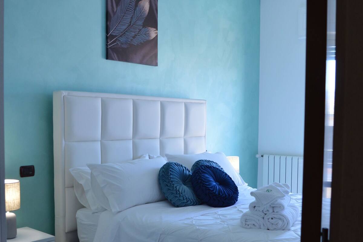 A neatly made bed with white bedding and blue pillows, a white nightstand with a lamp, and a wall art piece above the bed.