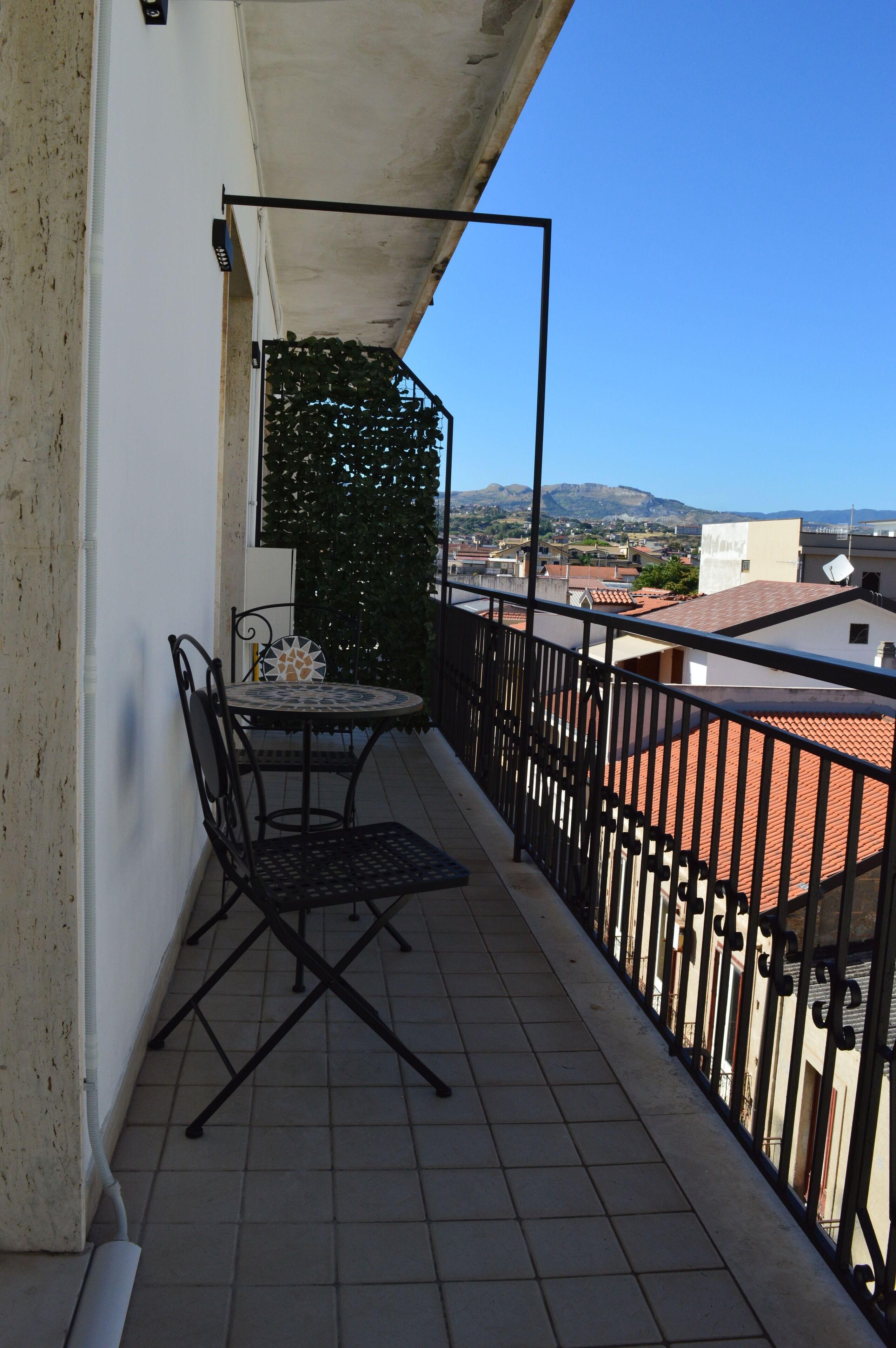 Un balcón con una mesa y sillas, vista a edificios y montañas, y un cielo azul despejado.