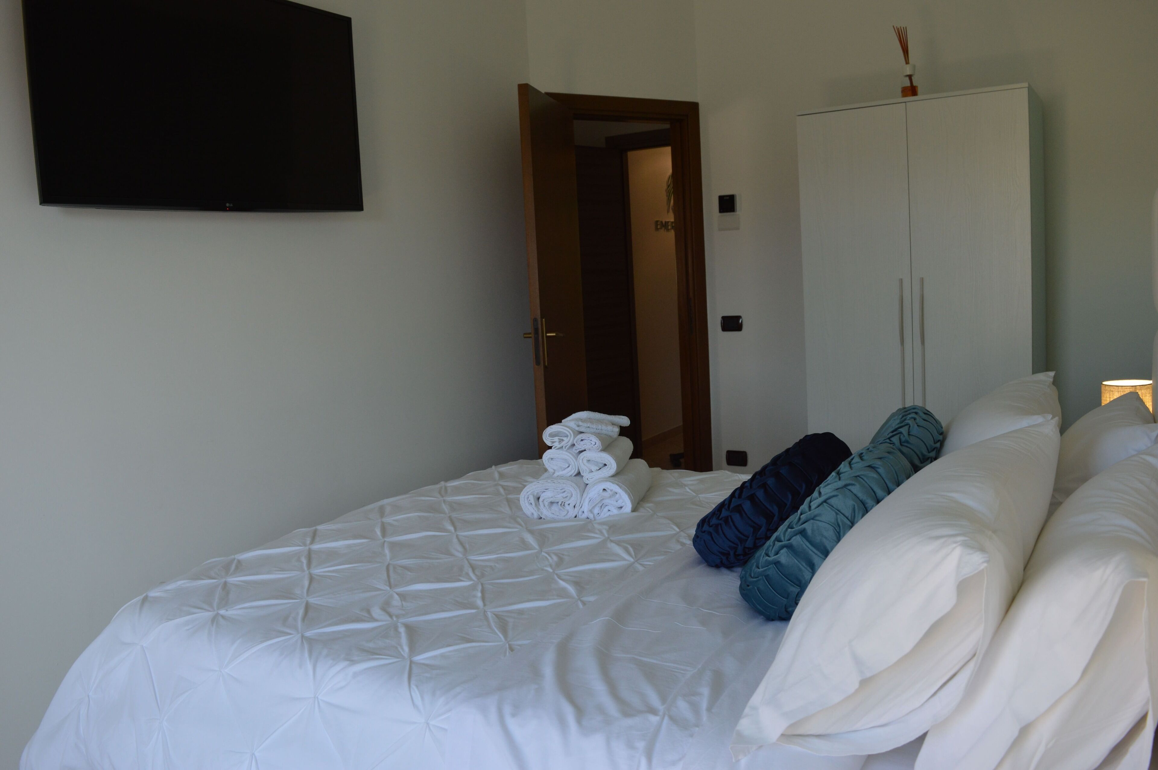 A neatly made bed with white and blue pillows, a rolled-up towel, and a white wardrobe with a small item on top.