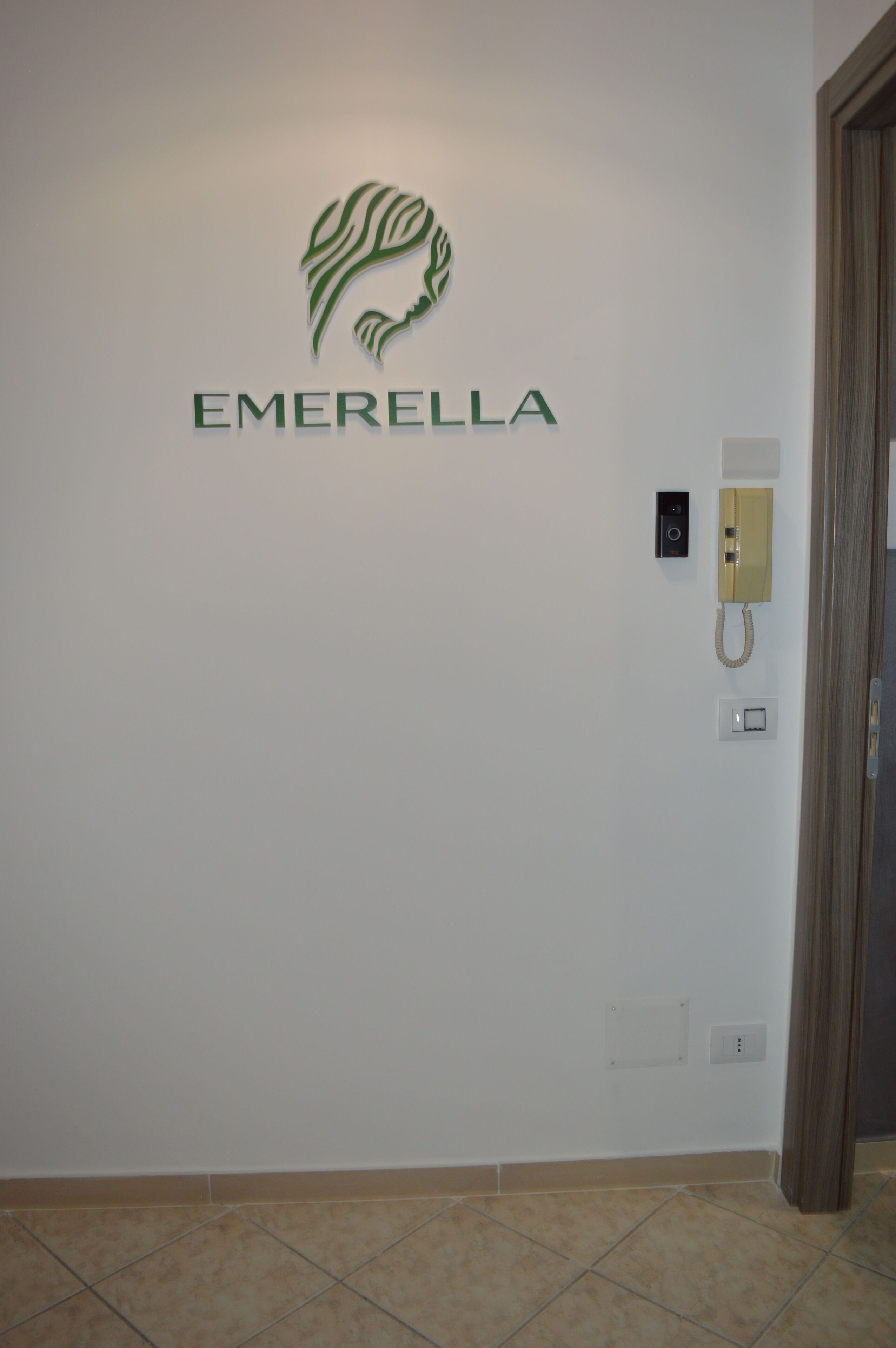 A wall with a logo and the name 'EMERELLA' in a hallway with a telephone and light switch.