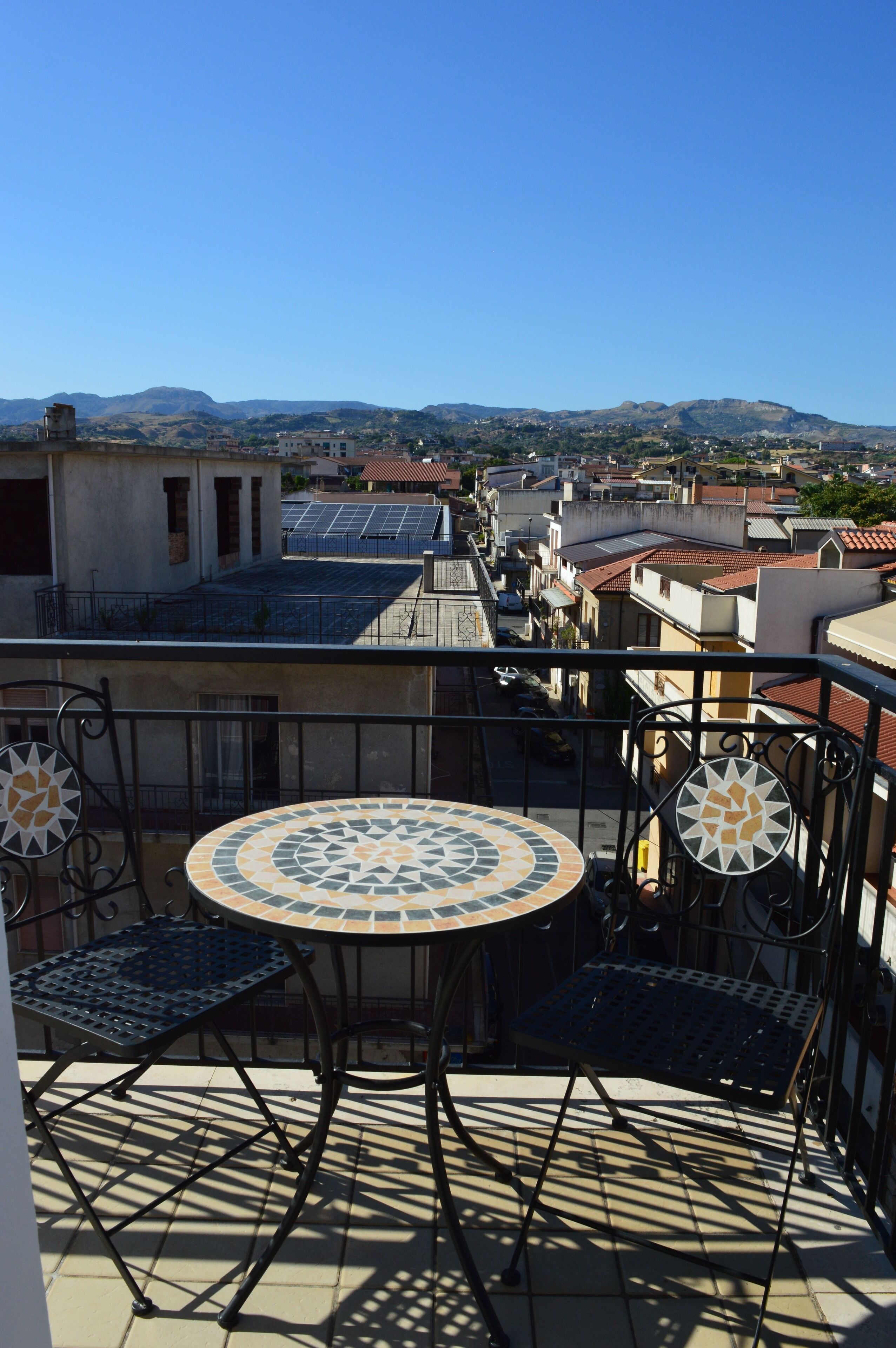 A balcony with a tiled table and chairs, overlooking a cityscape with buildings and mountains.