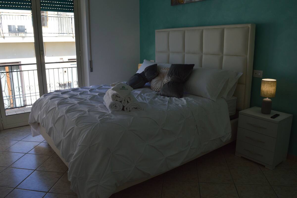 A bedroom with a bed, pillows, a nightstand, and a view of the outside balcony.