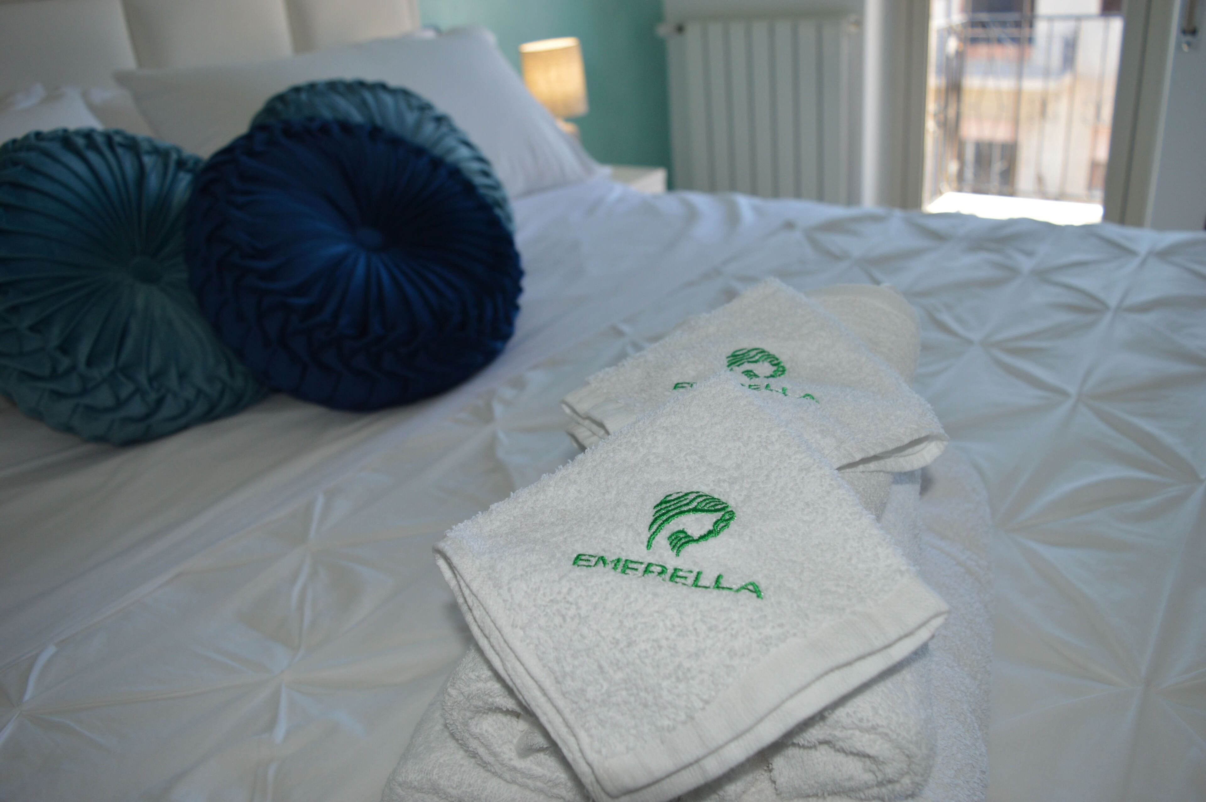 Una cama bien hecha con ropa de cama blanca y dos almohadas decorativas, una toalla doblada con un logo y una ventana con vistas a edificios.