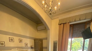 Una camera d'albergo con pavimento a scacchi, un letto con testiera, una scrivania con sedia, un lampadario e una finestra con tende.