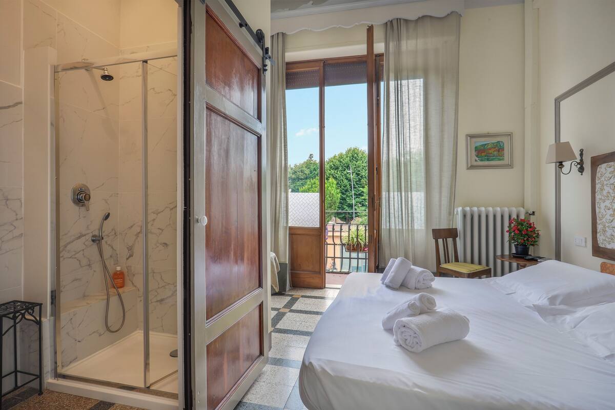 Habitación de hotel con una cama grande, ducha, balcón con vistas a árboles y una pequeña mesa con una planta.
