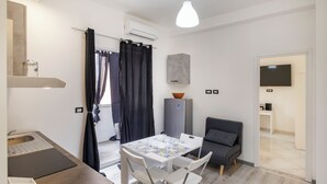 Un espacio habitable compacto con kitchenette, una zona de comedor con mesa y sillas, un sofá gris y un televisor montado en la pared.