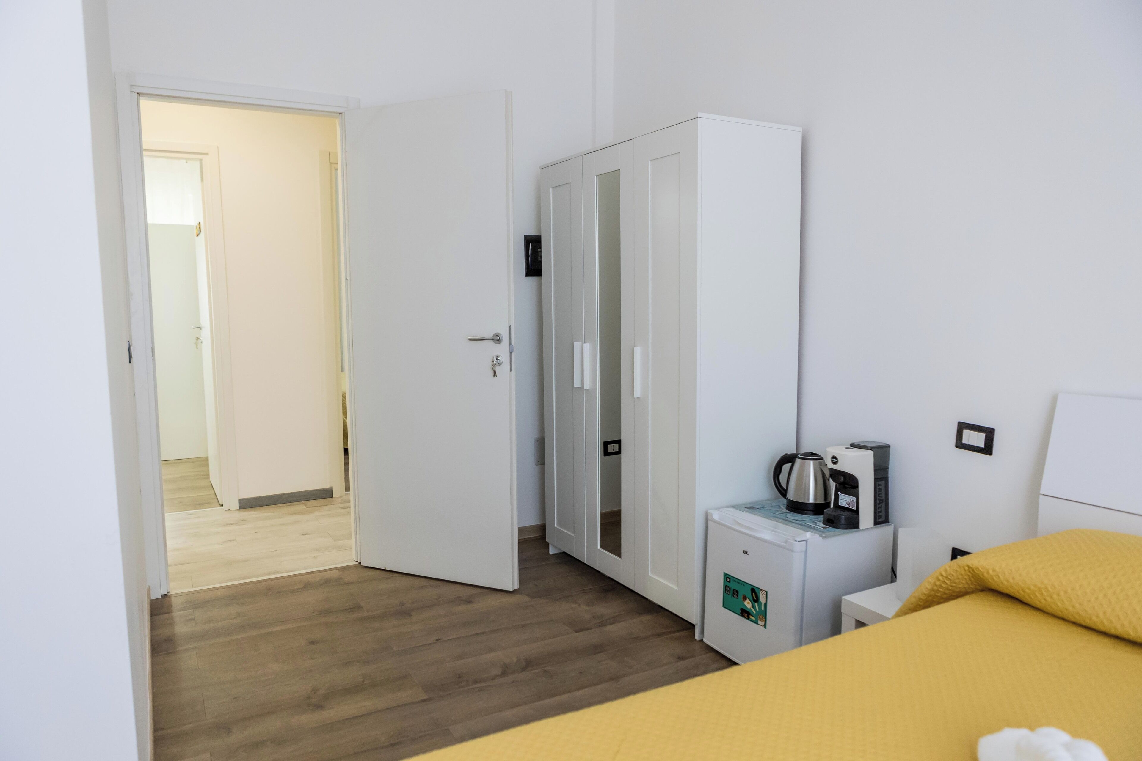 Una habitación de hotel con cama, armario, una pequeña kitchenette con cafetera y una puerta que da a otra habitación.