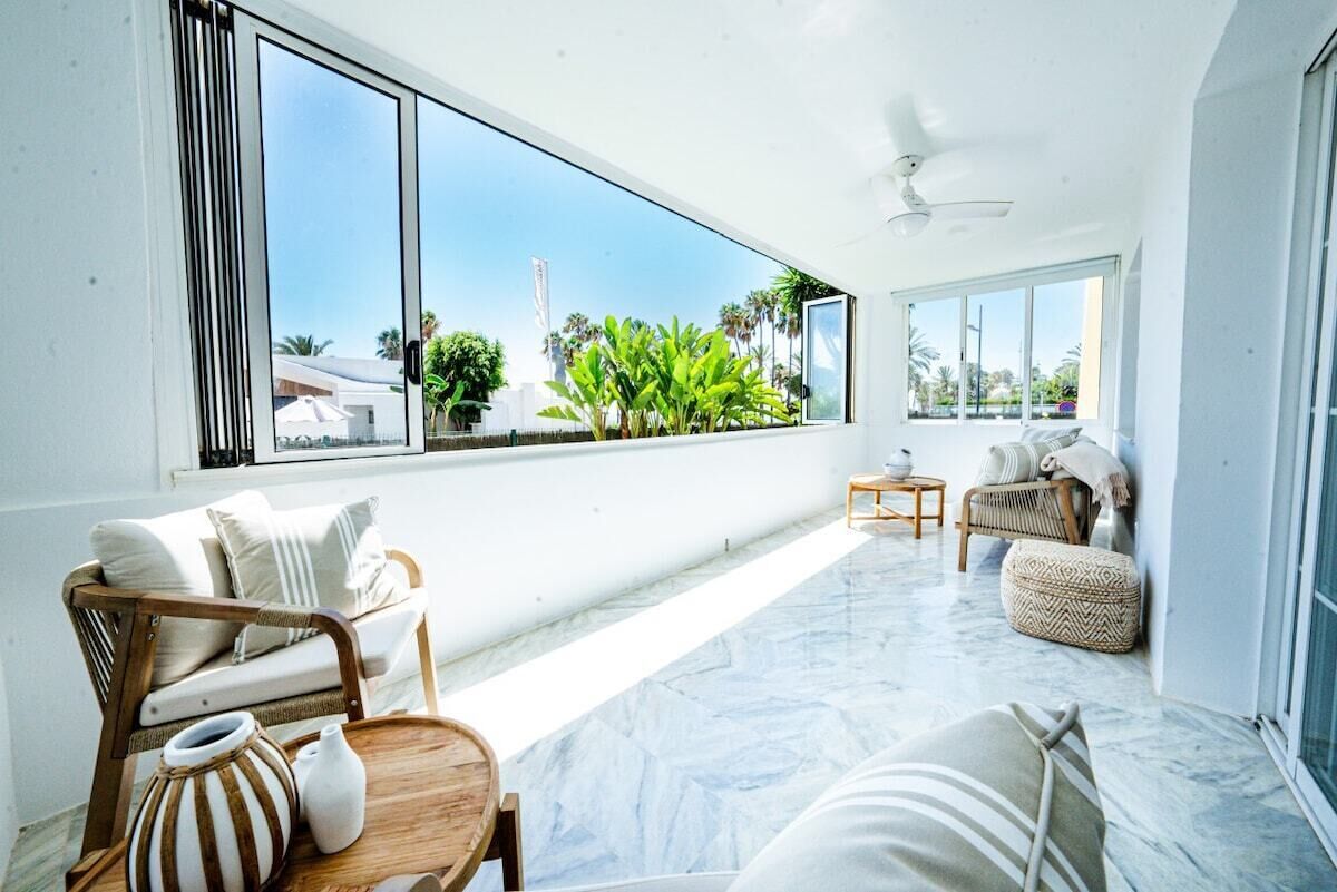 A spacious balcony with a view of palm trees and a building, furnished with a wicker chair, a small table, and a cushioned ottoman.