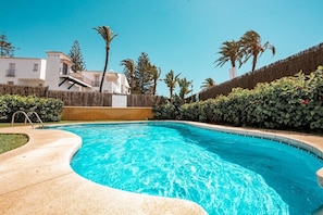 A swimming pool with a curved edge, surrounded by a low hedge, and a clear blue sky above.