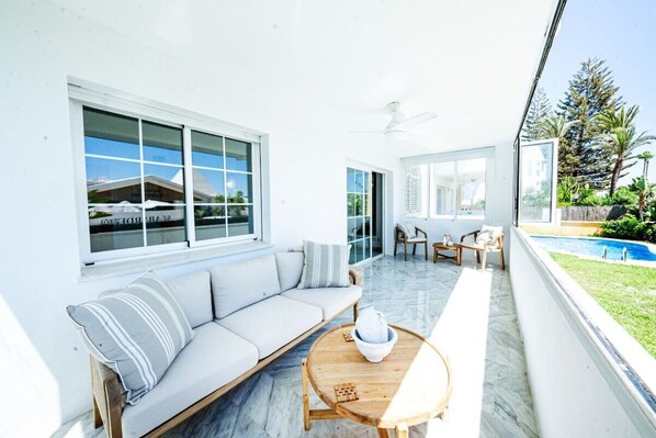 A bright, sunlit room with a white sofa, a wooden coffee table, and a view of a swimming pool.