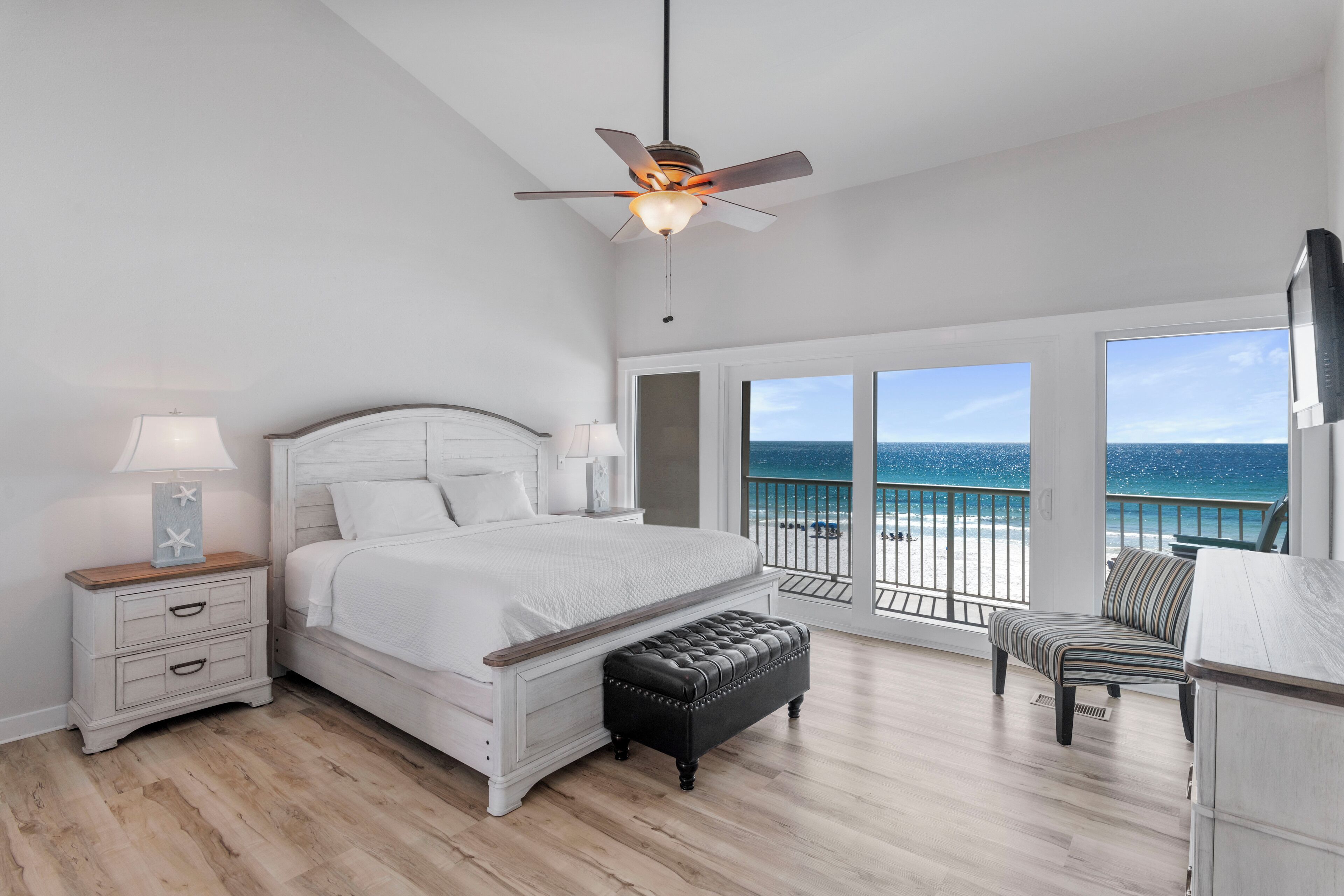 A bedroom with a bed, nightstand, bench, and a view of the ocean.