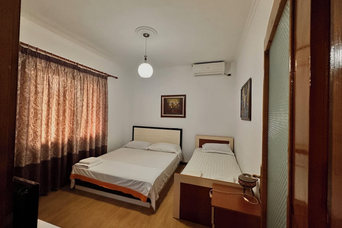 Ein Schlafzimmer mit zwei Betten, einem Deckenventilator, einer Klimaanlage und einem Fenster mit Vorhängen.