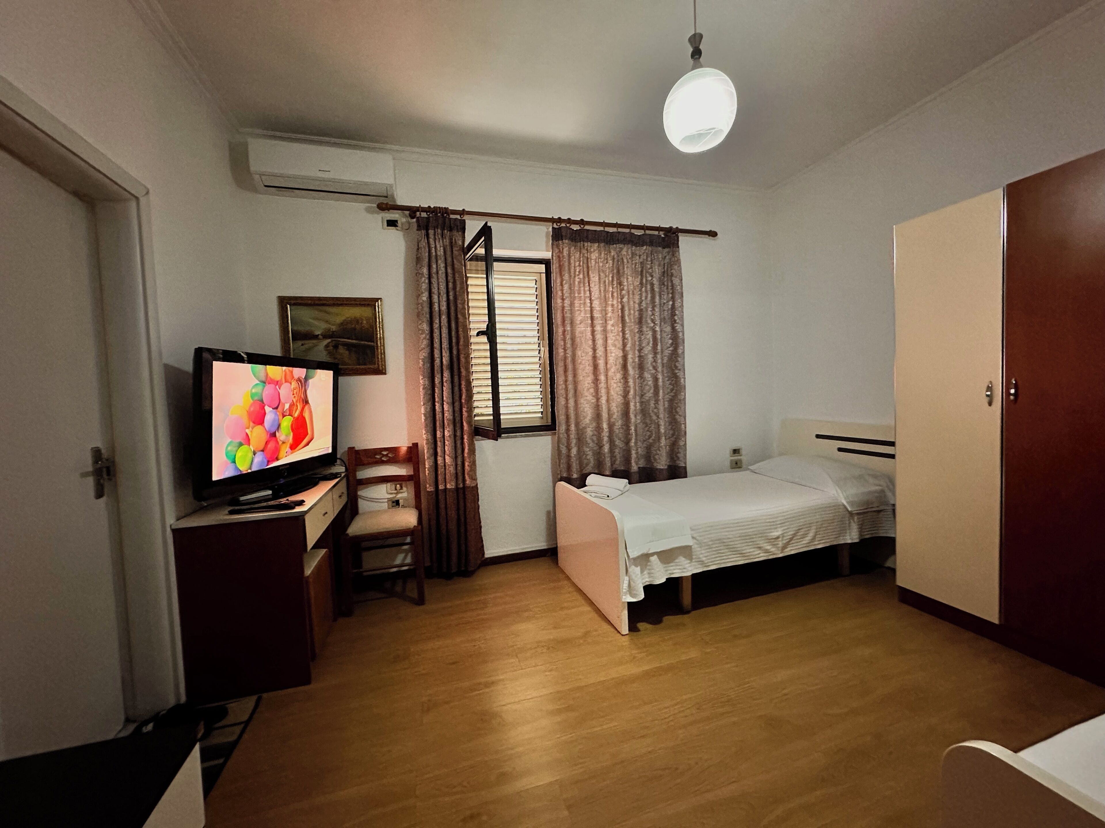 A small, well-lit room with a bed, a desk with a television, a chair, and a wardrobe.