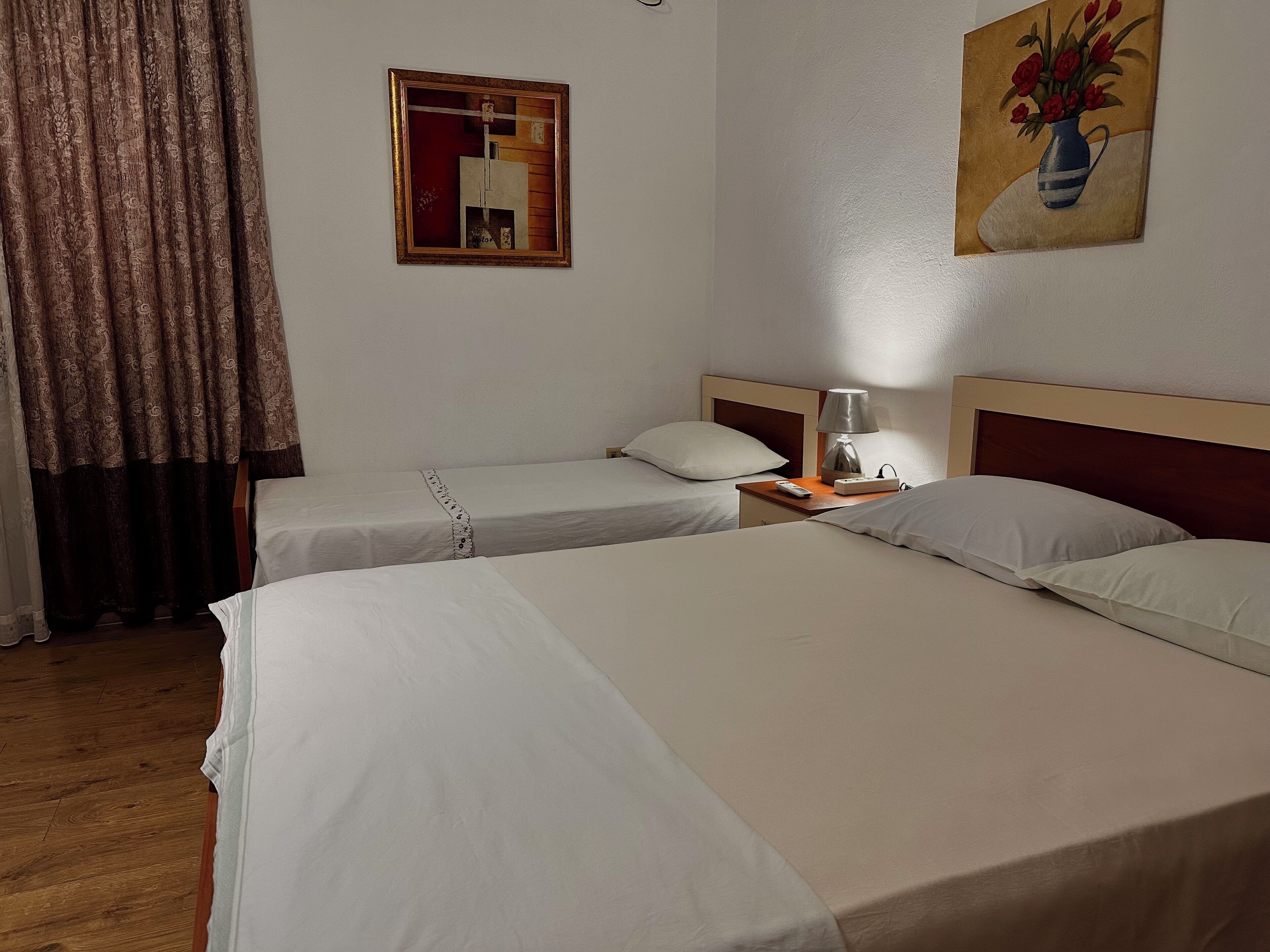 A hotel room with two beds, a nightstand with a lamp, a painting on the wall, and a curtain.