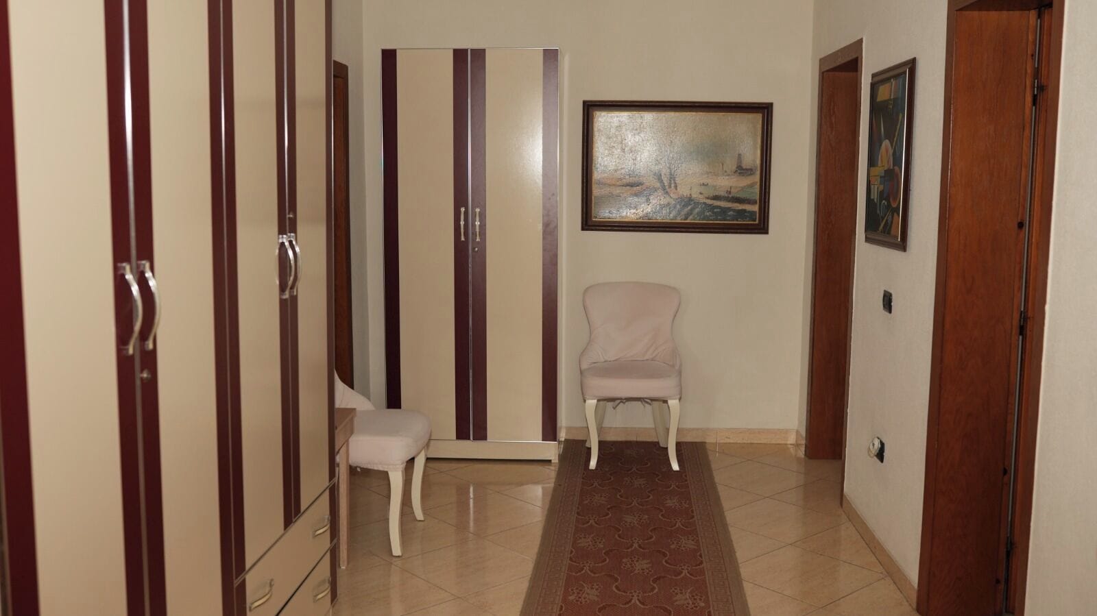A hallway with a wooden wardrobe, a white chair, a patterned rug, and a painting on the wall.