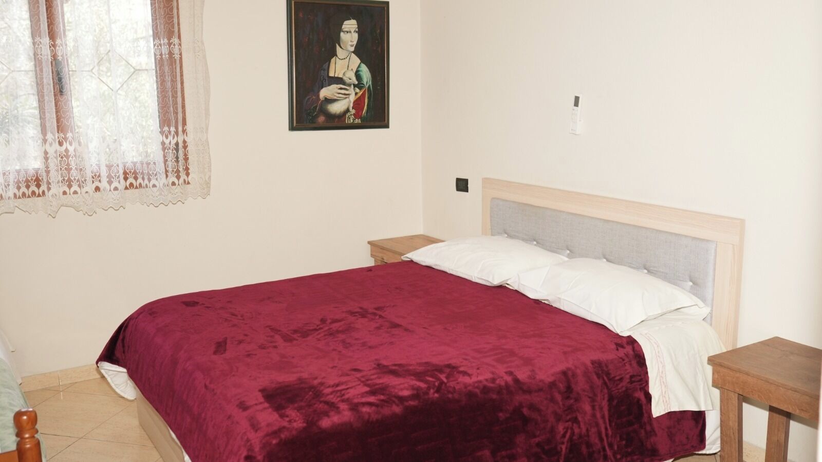 Un dormitorio con una cama grande, dos mesitas de noche, una mesita auxiliar, una ventana con cortinas de encaje y un cuadro en la pared.
