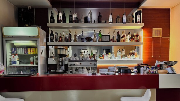 A bar area with shelves of liquor bottles, a coffee machine, and bar stools. - Grand House  (Tirana)