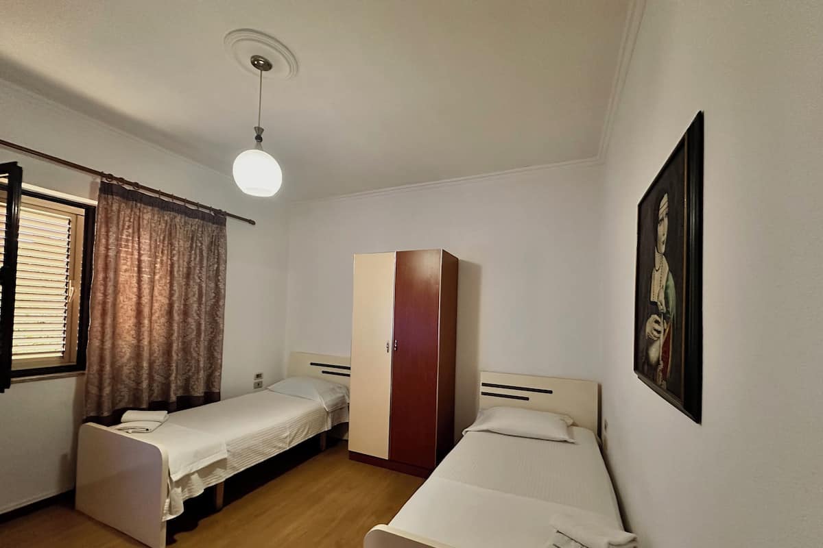 Ein kleines, sauberes Zimmer mit zwei Einzelbetten, einem Kleiderschrank, einer Deckenleuchte und einem Fenster mit Vorhängen.