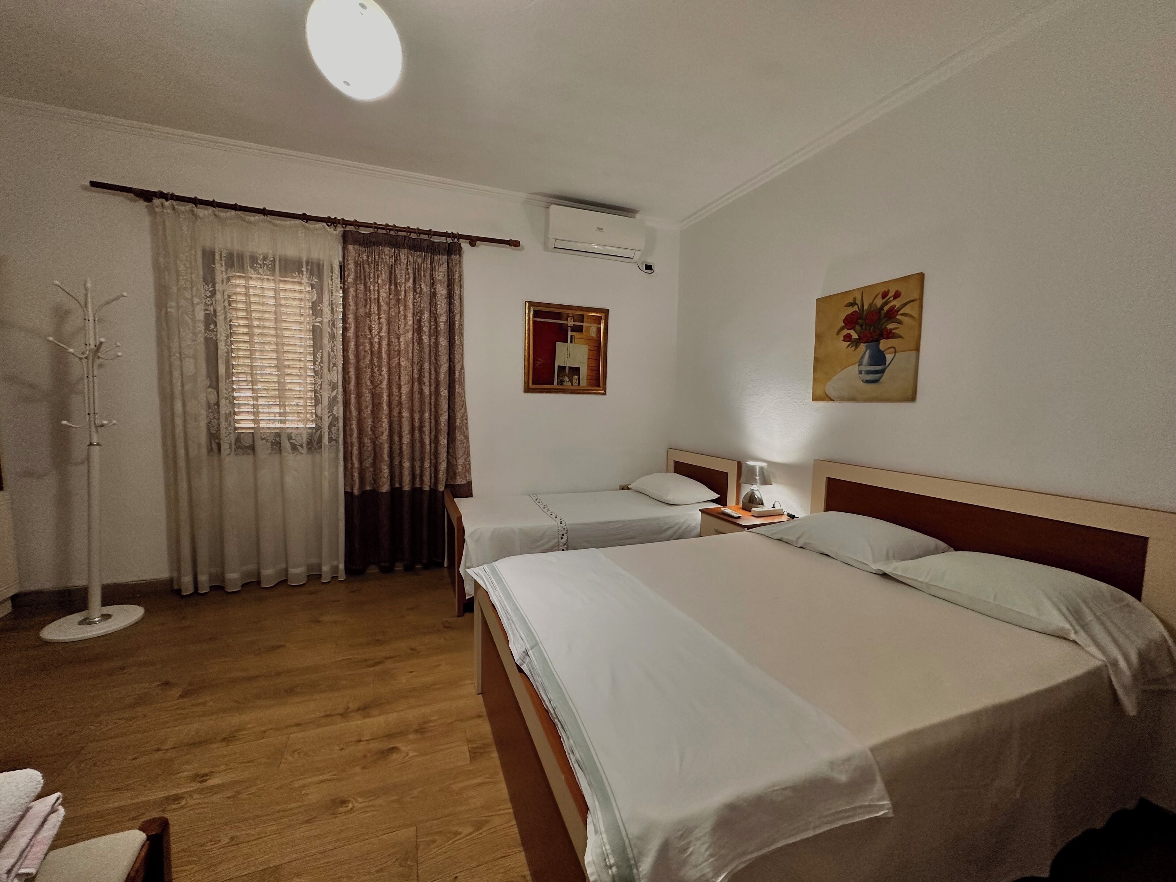 A hotel room with two beds, a coat rack, a window with curtains, an air conditioning unit, and a painting on the wall.