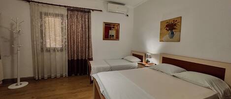A hotel room with two beds, a coat rack, a window with curtains, an air conditioning unit, and a painting on the wall.