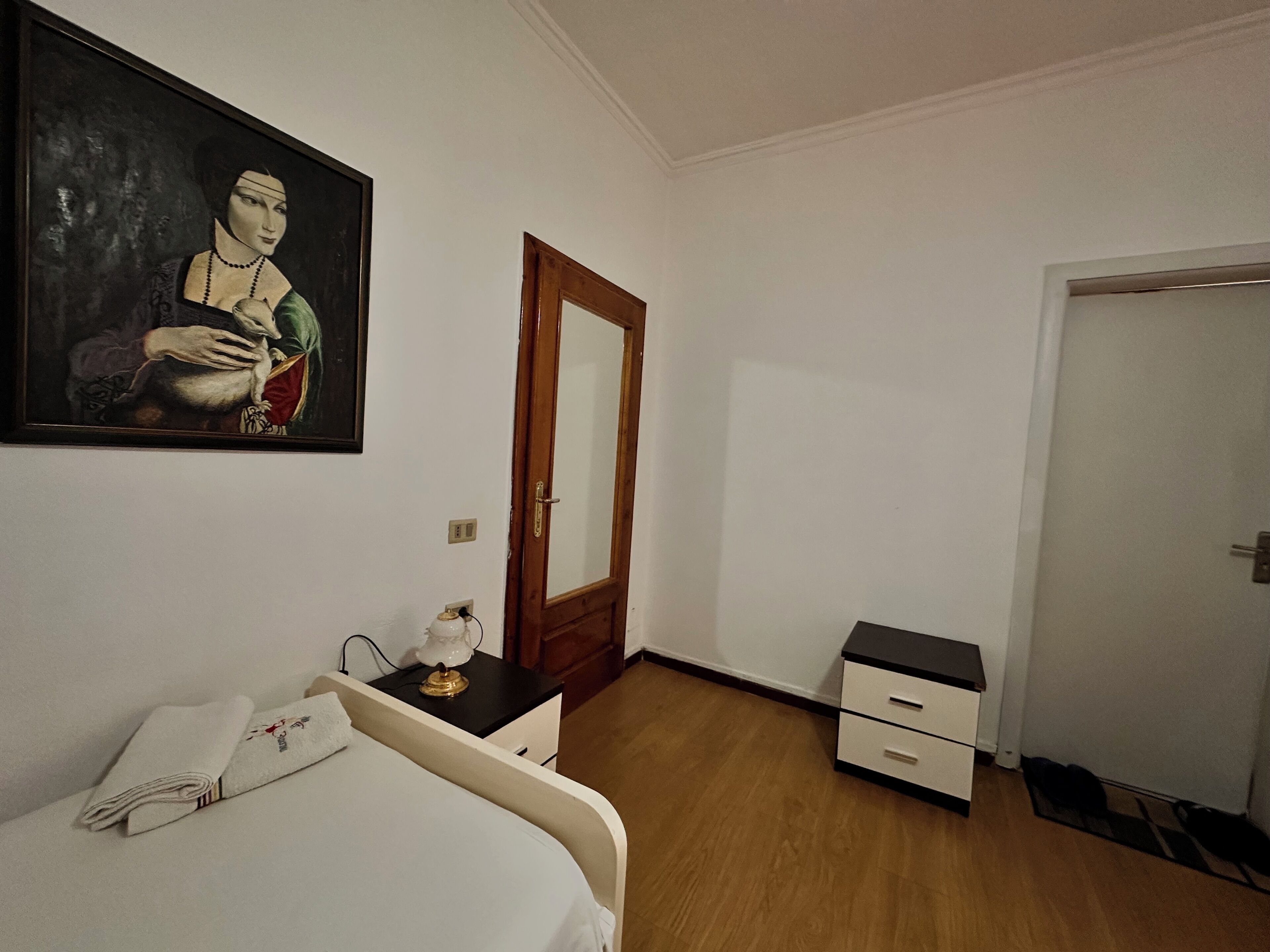 Um quarto com uma cama, uma mesinha de cabeceira com luminária, um armário pequeno, uma porta e um quadro na parede.