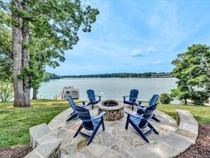 An outdoor seating area with Adirondack chairs and a stone fire pit, overlooking a lake with a dock and a small building. - 16' of Water @ Full Pond!  <br>3 Bedrooms, 1 Flex Room, 3½ Bathrooms!  Come Visit FOREVER YOUNG! (Moneta)