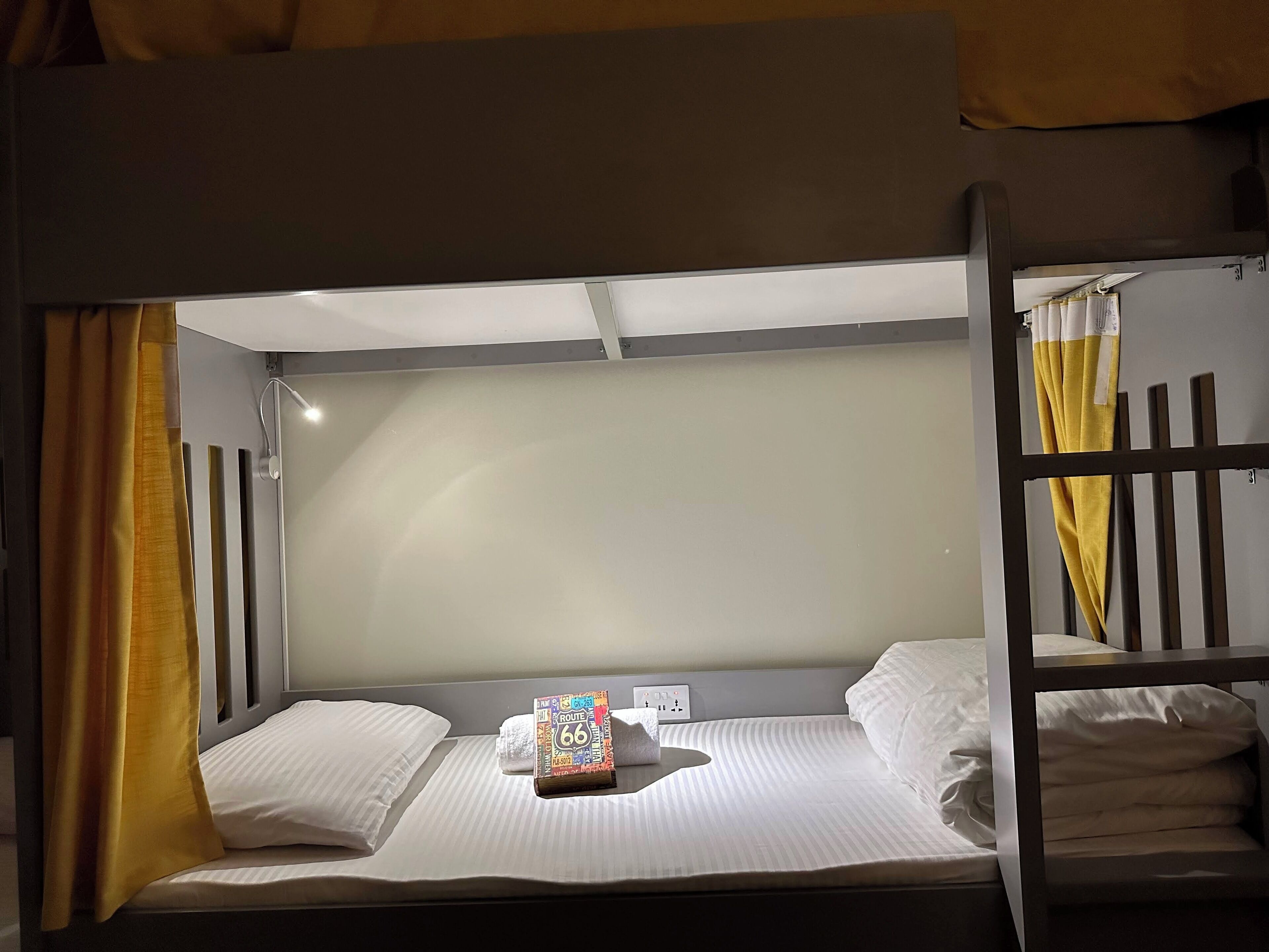 A bunk bed room with a single bed at the bottom and a headboard with a light fixture. There is a small table with a book and a tissue box on the bed.
