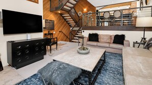Una sala de estar moderna con un televisor de pantalla plana grande, un mueble de entretenimiento negro, un sofá color beige, una mesa de centro y una escalera con interior de madera.