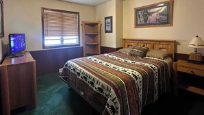 Un dormitorio con cama, escritorio con computadora, ventana con persianas, estante y un cuadro en la pared.