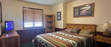 Un dormitorio con cama, escritorio con computadora, ventana con persianas, estante y un cuadro en la pared.