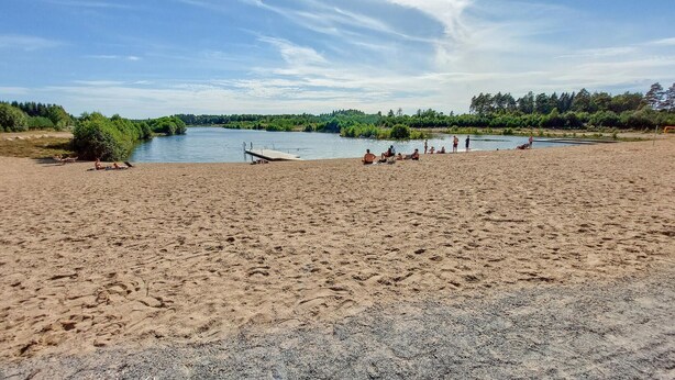 Ein Sandstrand mit einem Steg, der in einen ruhigen, von Grün umgebenen See führt.