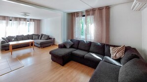 Ein modernes Wohnzimmer mit einer Eckcouch, einem Holztisch und einem großen Fenster mit transparenten Vorhängen.