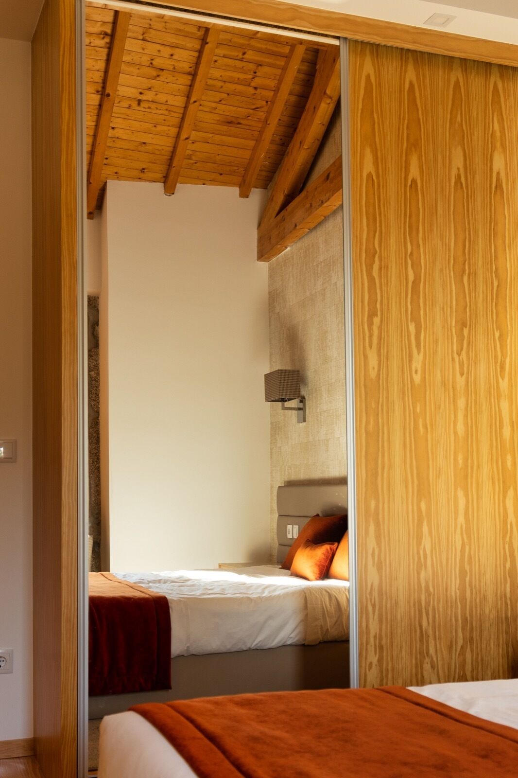 A bedroom with a wooden ceiling, a bed with white bedding and orange pillows, and a wall-mounted light fixture.