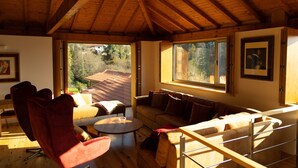 A cozy living room with a wooden ceiling, a sofa, a red armchair, a small round table, and large windows offering a view of a house and trees. - Casa S. Miguel Arcanjo - T5 (Caniçada)