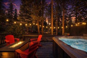 Un bain à remous entouré de chaises rouges sur une terrasse en bois, avec des guirlandes lumineuses et une forêt en arrière-plan.
