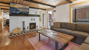 A cozy living room with a fireplace, wooden ceiling, and a large window. - Navigate Stays Hot Tub Home 5 Min to Ski Resort (Big Bear Lake)