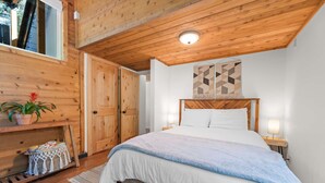A cozy room with a wooden ceiling, a bed with white linens, a nightstand with a lamp, and a wall-mounted geometric tapestry. - Navigate Stays Hot Tub Home 5 Min to Ski Resort (Big Bear Lake)