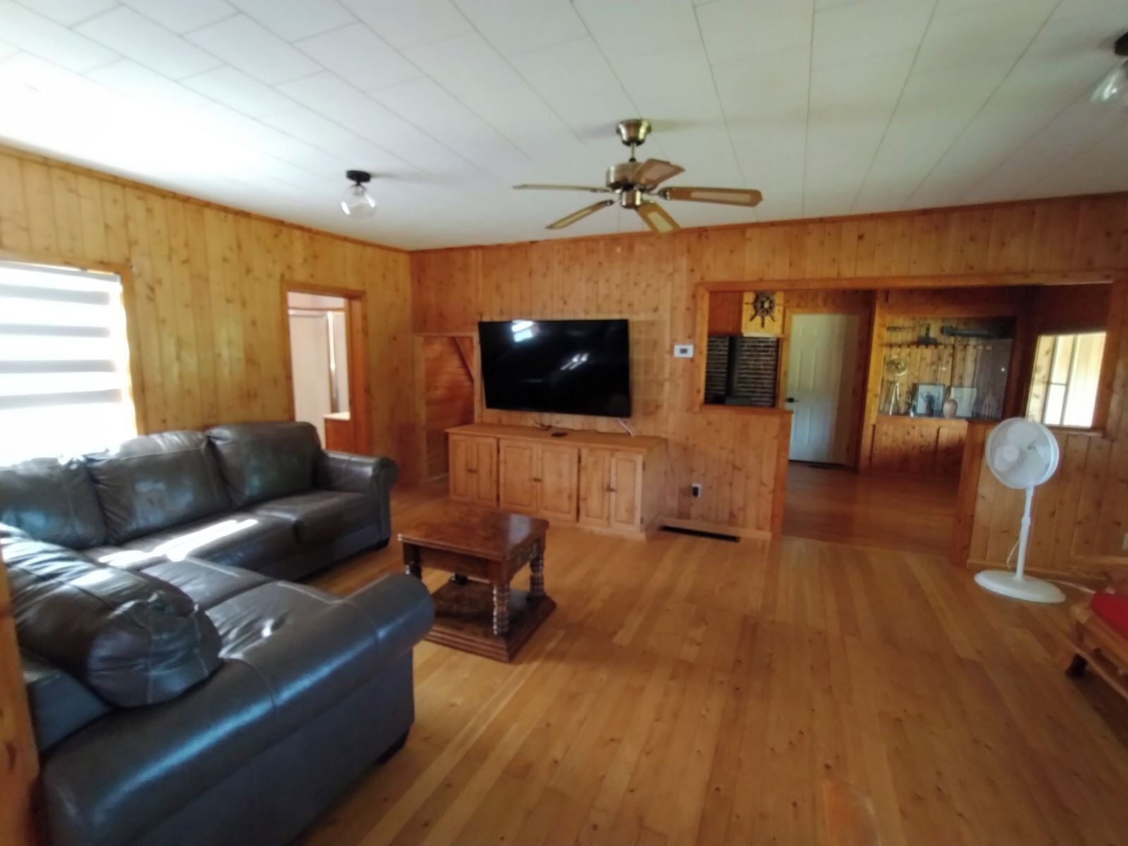 A wooden-paneled living room with a leather sofa, a television, and a ceiling fan.