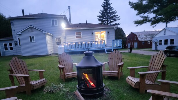 Een achtertuin met een vuurkuil, omringd door houten stoelen, een huis op de achtergrond en een veranda met verlichting.