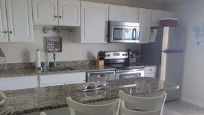 Cocina con armarios blancos, encimera de granito, refrigerador de acero inoxidable y microondas. Hay tres taburetes blancos con cojines marrones.