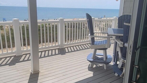 Una terraza cubierta al aire libre con vista a la playa, barandilla blanca y una mesa con sillas.