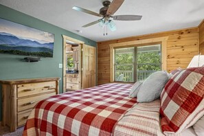 Un dormitorio con colcha a cuadros, cómoda de madera, ventilador de techo, ventana con persianas y un cuadro en la pared.