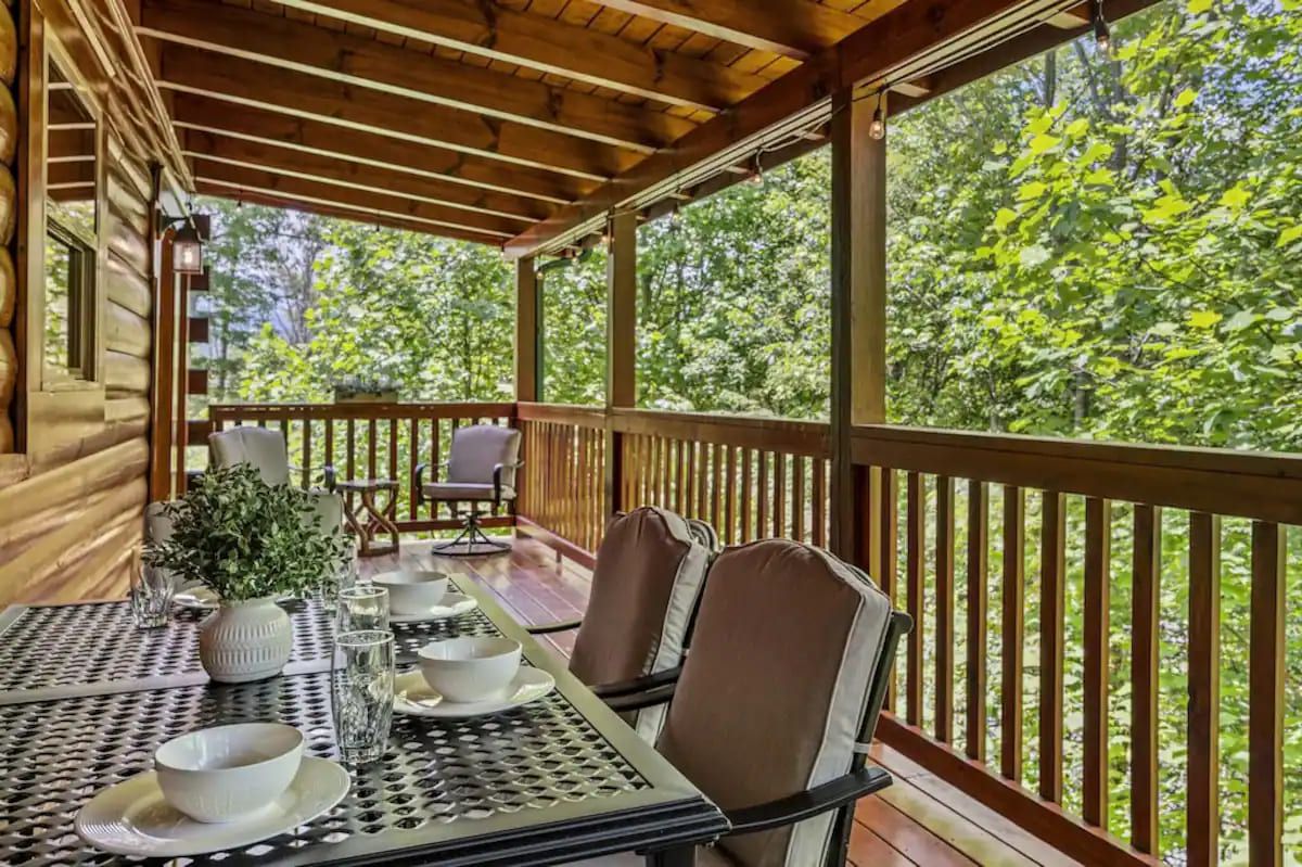 Un espacio exterior techado para comer con una mesa preparada para dos personas, rodeada de sillas, con vistas a una zona boscosa.