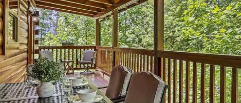 Un espacio exterior techado para comer con una mesa preparada para dos personas, rodeada de sillas, con vistas a una zona boscosa.
