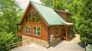 A log cabin with a stone foundation, a green metal roof, and a porch with a railing.