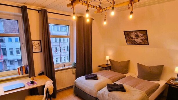 Une chambre d’hôtel avec deux lits, un bureau et une chaise. Il y a une fenêtre avec des rideaux, un radiateur et un tableau au mur.