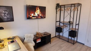 Un salon moderne avec un téléviseur à écran plat, une table basse en bois, un fauteuil moelleux, un meuble TV noir avec un plateau, et une étagère en métal où sont suspendus des vêtements.